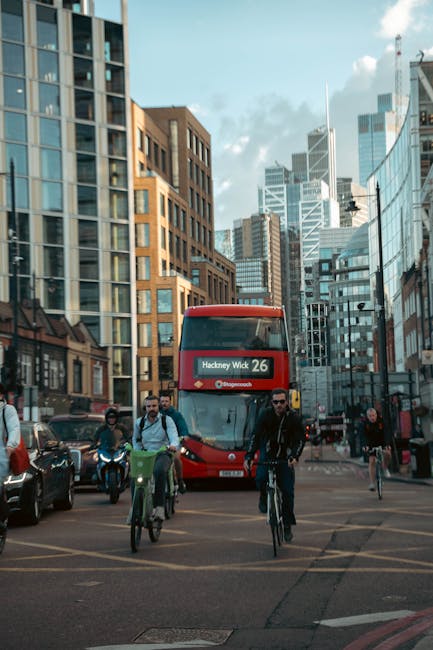 A busy city street scene in Hackney with a red double-decker bus displaying 'Hackney Wick 26' approaching an intersection amidst tall modern office buildings made of glass and brick. Several pedestrians and cyclists are crossing the street, with some waiting on the sidewalk. The scene is illuminated by daylight with partly cloudy skies overhead. This urban environment reflects typical city traffic and movement, relevant to local house removal and furniture transport services. Hackney Man and Van may assist with home relocation or moving logistics, ensuring smooth handling of furniture, boxes, and packing materials during the loading process amid city traffic.