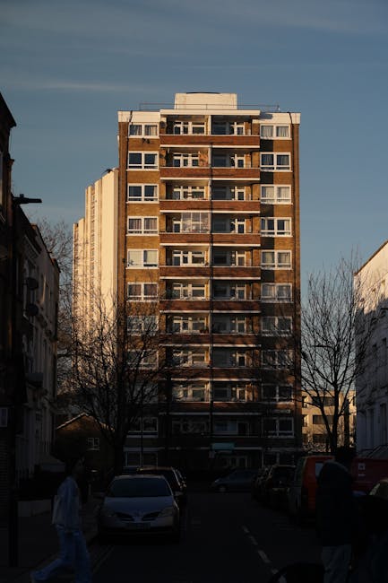 A multi-storey residential building with a brick facade and numerous balconies, situated in an urban area with other smaller buildings on either side. The building towers over a parking area at street level, where several cars are parked and a few pedestrians are walking nearby. Leafless trees line the street, and the sky above is partly cloudy with patches of blue. The scene appears to be captured during late afternoon, with natural light casting shadows on the building. This image reflects a typical scene in preparation for home relocation or furniture transport, as part of a house removal process. Hackney Man and Van offers professional moving and packing services for flats and apartments, supporting efficient logistics of house removals in Hackney, particularly around Dalston's Mare Street E8 area.