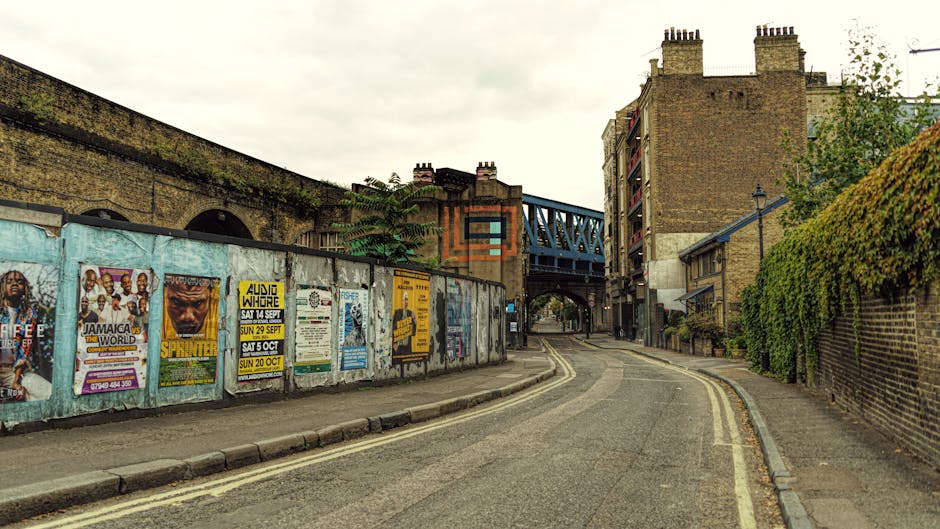 A narrow street in Hackney, London, featuring a row of posters attached to a weathered metal fence on the left side, advertising events such as reggae concerts and local performances. The street is paved with asphalt and has double yellow lines along the curb, indicating parking restrictions. On the right, there is a brick wall partially covered with green ivy and some sparse foliage, with residential buildings visible behind it. The scene includes a bridge overhead painted blue, connecting two buildings, with a small archway beneath it allowing vehicles to pass through. The sky is overcast, casting diffuse natural light over the urban environment. In the background, a van or truck is partially visible, hinting at the logistics involved in local furniture transportation and home relocation, with the overall setting consistent with a typical Hackney street scene associated with removals and moving services, such as those provided by Hackney Man and Van.