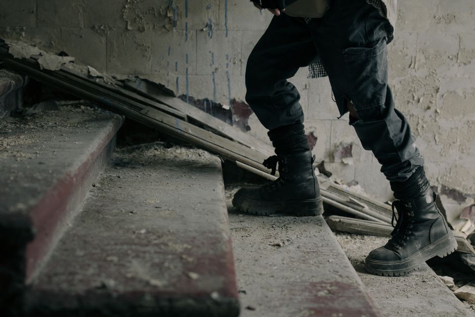 A person wearing black work boots, black cargo pants, and a black jacket is shown lifting and carrying stacked cardboard boxes on a home staircase. The staircase is made of concrete with visible dust, debris, and chipped paint along the edge. The background reveals a rough, unfinished wall with exposed cement and patches of plaster, indicating ongoing renovation or construction work. Nearby, a yellow van with an open rear door is visible, suggesting loading or unloading for a house removal or relocation process. The scene captures the challenging environment of moving furniture and boxes through difficult access points, supporting professional removal services like those offered by Hackney Man and Van during home relocations involving tight staircases or tenement buildings.