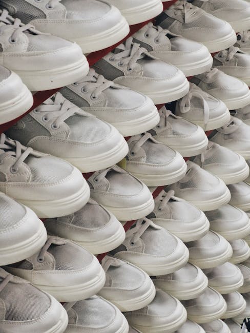 Close-up view of multiple pairs of white and grey athletic sneakers arranged in rows on a display shelf, with some sneakers overlapping each other. The sneakers are made of fabric and synthetic materials, with white laces and rubber soles. The background shows part of a retail store environment, highlighting the packaging and display arrangement. This visual is relevant to the process of packing or preparing footwear for a move or delivery, which is part of household relocation or home removals services offered by Hackney Man and Van, especially when handling delicate or bulkier items during a difficult access move involving a tenement staircase in Hackney.