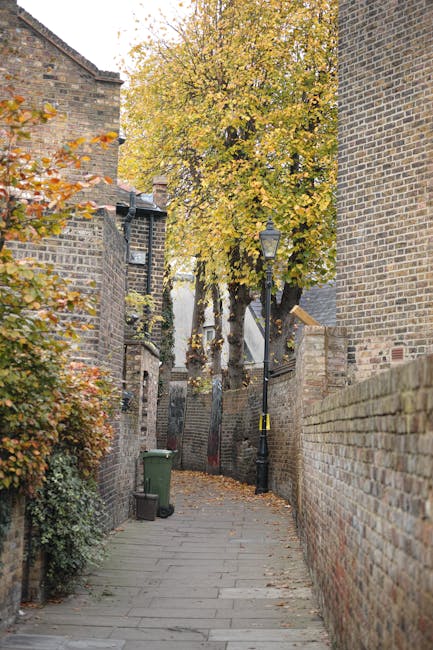 A narrow residential street with a paved walkway, flanked by tall brick walls on both sides. On the left, dense foliage with autumn-colored leaves extends upwards and partially covers parts of the walls, while on the right, a tall brick wall runs along the pavement. A vintage-style street lamp is mounted on a black pole attached to the brick wall on the right, illuminating the walkway. At the end of the alley, a large tree with yellow and orange leaves overhangs, with some leaves scattered on the ground. A green waste bin is positioned near the corner on the left side, and a black trolley is visible in the distance, suggesting recent or ongoing furniture or box loading activities associated with home relocation or moving services. The scene is outdoors, with natural daylight illuminating the area, reflecting a typical urban setting suitable for house removals or furniture transport, as seen in the Hackney Wick man and van guide.