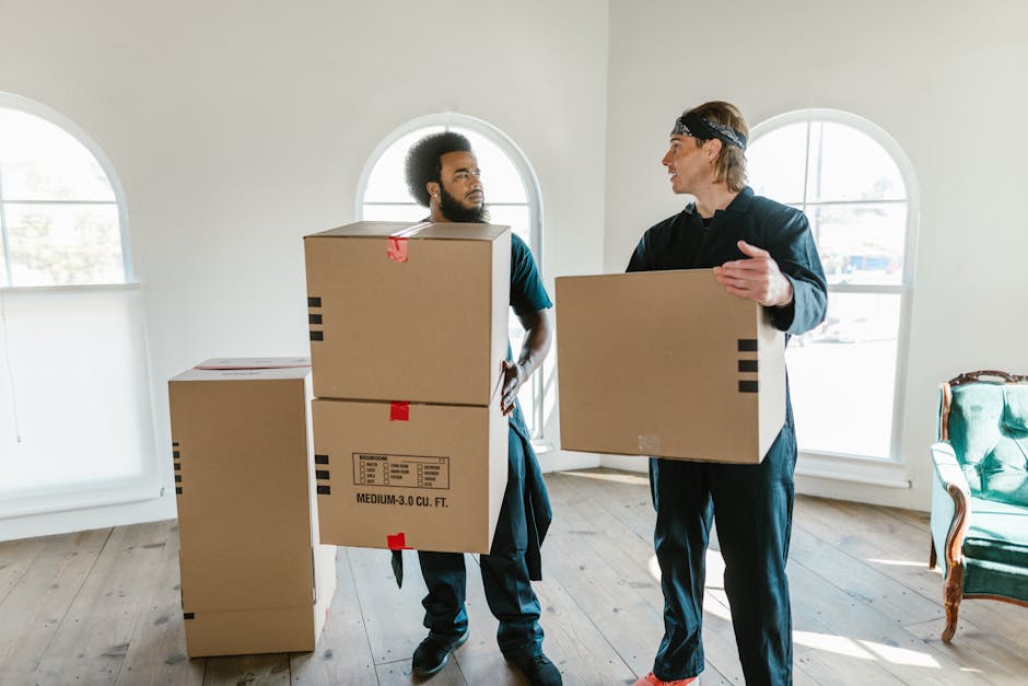 Two professional movers from Hackney Man and Van are inside a well-lit room with white walls and large arched windows. One mover, with curly dark hair and a beard, is carrying a medium-sized cardboard box with red tape along the top and bottom edges, standing near a stack of similar boxes on the floor. The second mover, with blonde hair and wearing a headband, is holding a larger cardboard box and engaged in conversation with the first. Several more cardboard boxes, some sealed with tape, are arranged on the wooden floor near the window, ready for home relocation. The room contains minimal furniture, including a green upholstered armchair visible on the corner, and the scene appears to be part of a packing and moving process in preparation for furniture transport or local removal services. The light streaming through the windows provides natural illumination, emphasizing the careful handling of items during packing and loading for a house removal.