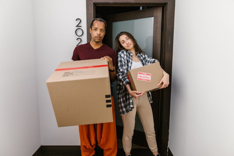 A scene featuring a variety of tropical and subtropical plants, including a large, weathered palm tree with some brown, drooping fronds, and smaller green palm-like plants with spiky leaves in the foreground. Behind the plants, there is a collection of residential buildings with light-colored facades, some with balconies, and rooftops in shades of red and beige. In the background, a prominent church tower with a clock and a bell, painted in yellow and brown, rises above the buildings, set against a backdrop of lush green mountains under a partly cloudy sky with patches of blue. The image captures an outdoor suburban or small-town environment, with the tree and plants situated on a patch of garden or park area adjacent to the houses. The scene suggests a warm climate, typical of Mediterranean or similar regions, relating to home relocation or packing and moving procedures in such areas, with Hackney Man and Van providing professional removals services that may include transport to similar environments.