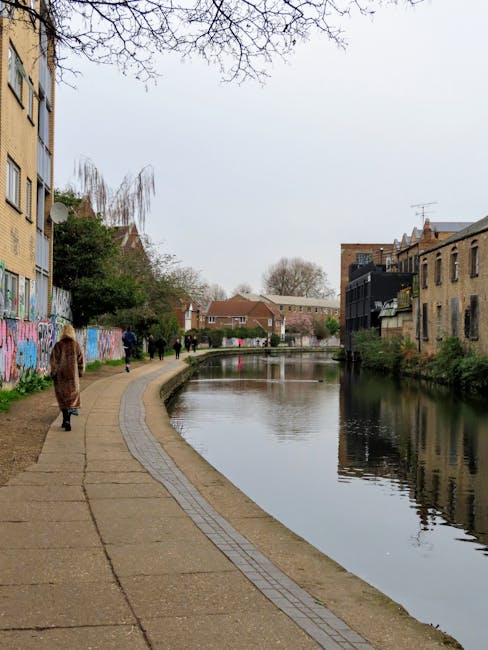 A paved walkway runs alongside a narrow river or canal in an urban area during daytime, with several people walking along the path. To the left, there are multistory residential buildings with yellow